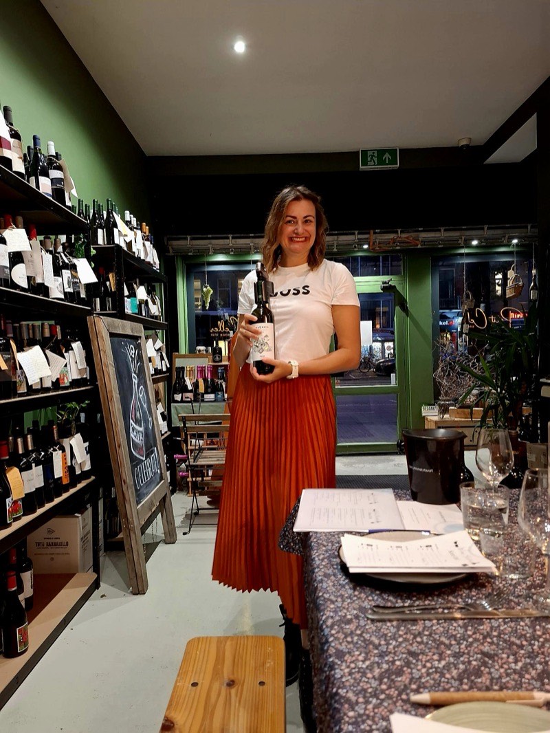 Biljana met een fles wijn tussen de schappen van een wijnwinkel