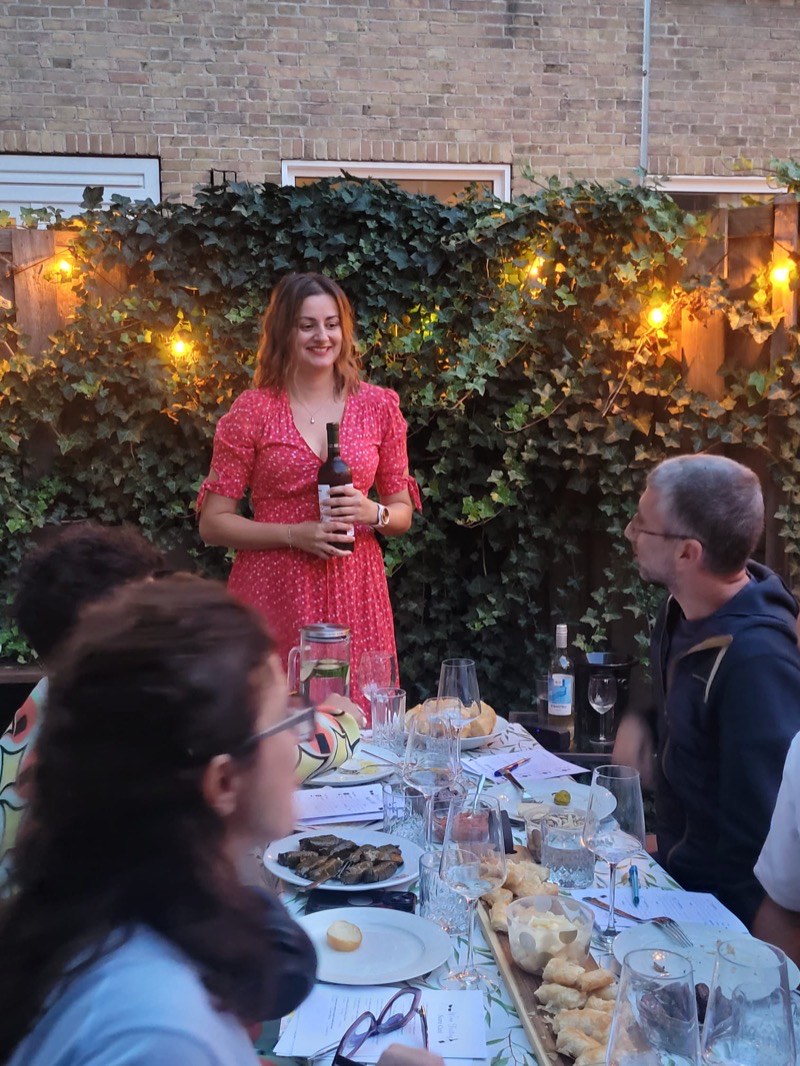 Biljana vertelt over een wijn aan een tafel buiten, tussen klimop en lichtjes